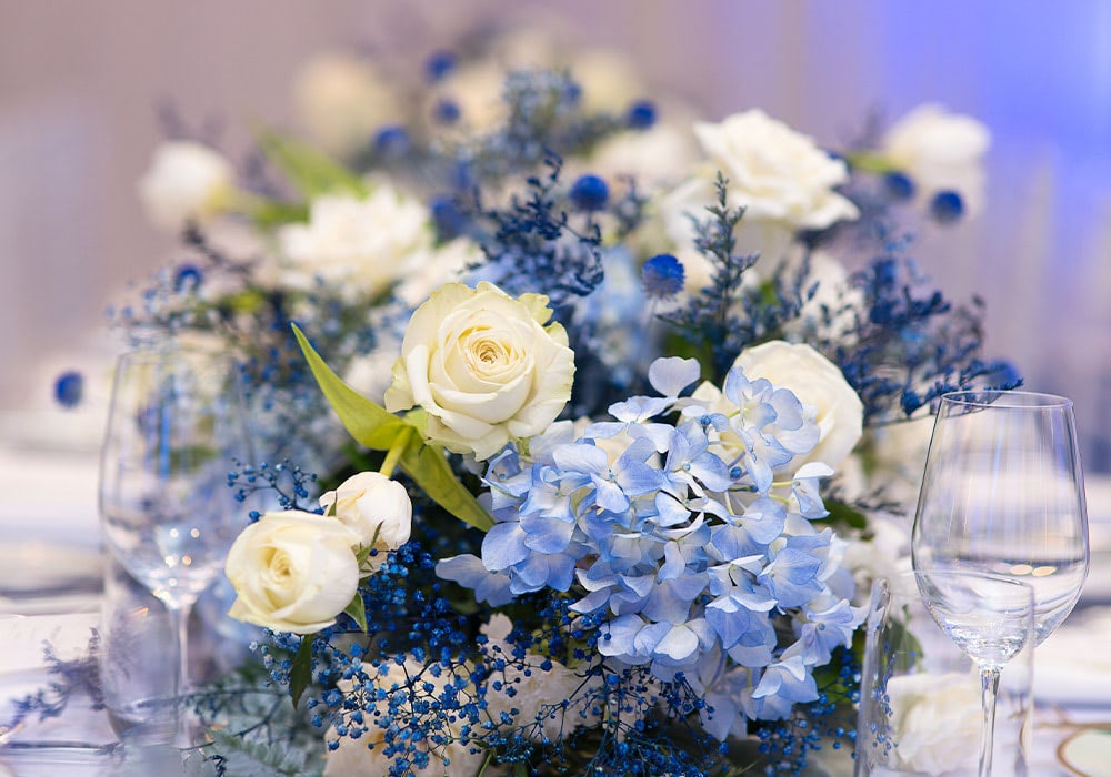 Bouquet of white roses, blue hydrangeas, and greenery arranged on a table surrounded by empty wine glasses, creating an elegant and sophisticated centerpiece in a softly lit setting.