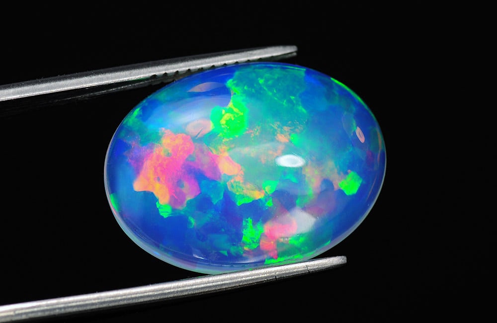 A vibrant, multi-colored opal gemstone, held by silver tweezers against a black background, displays hues of blue, green, pink, and yellow.